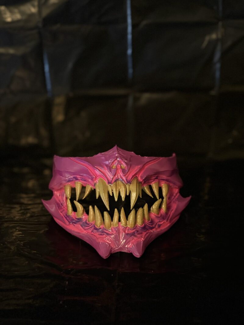 3D printed demon jaw mask with sharp golden fangs and pink creature design.